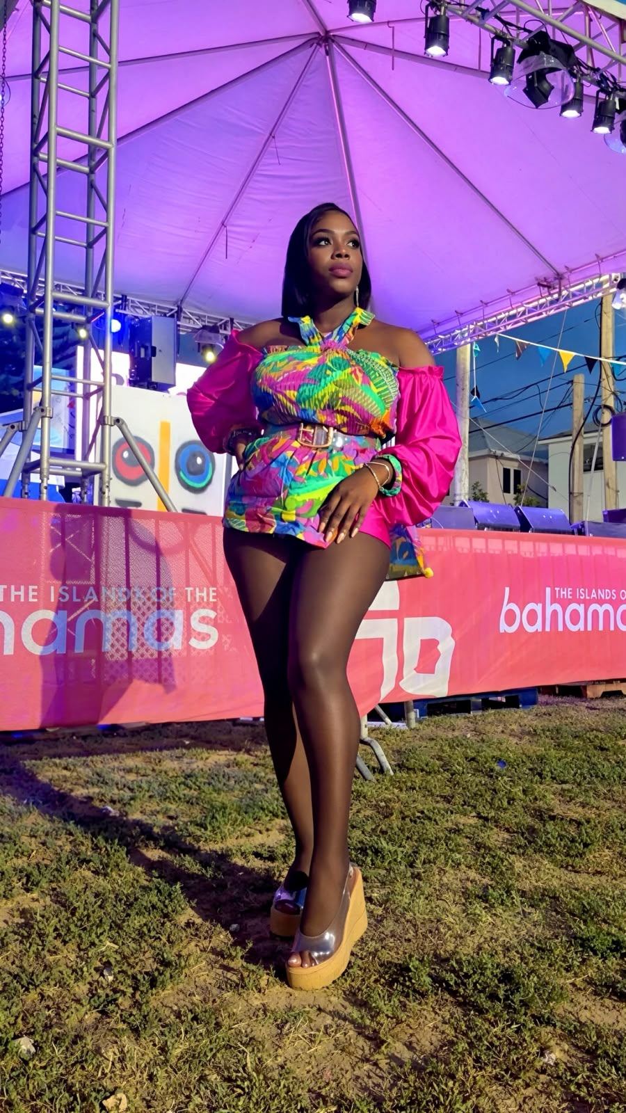 Mama D in a multicoloured printed outfit with hot pink sleeves standing in front of an Islands of The Bahamas banner