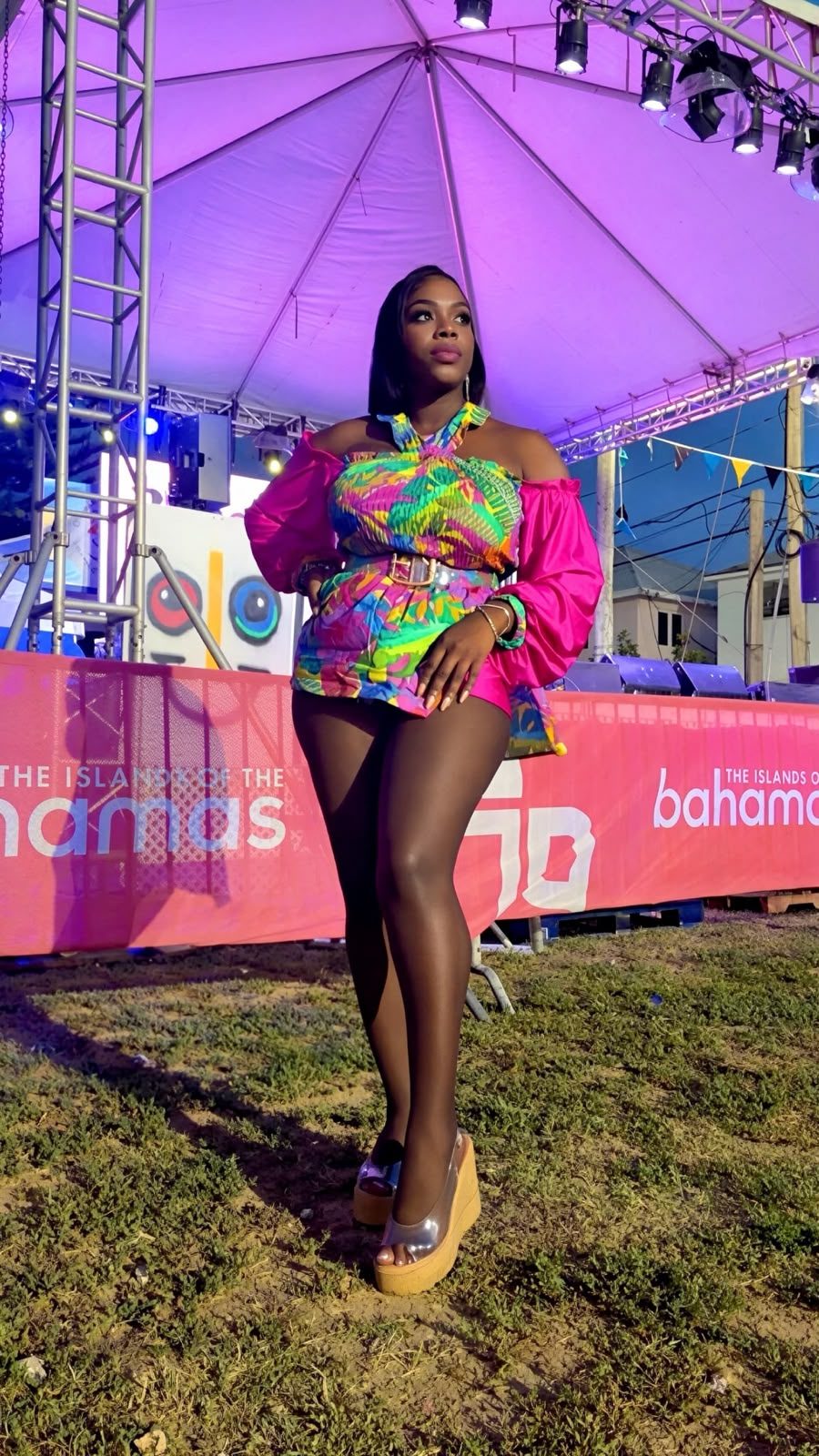 Mama D in a multicoloured printed outfit with hot pink sleeves standing in front of an Islands of The Bahamas banner