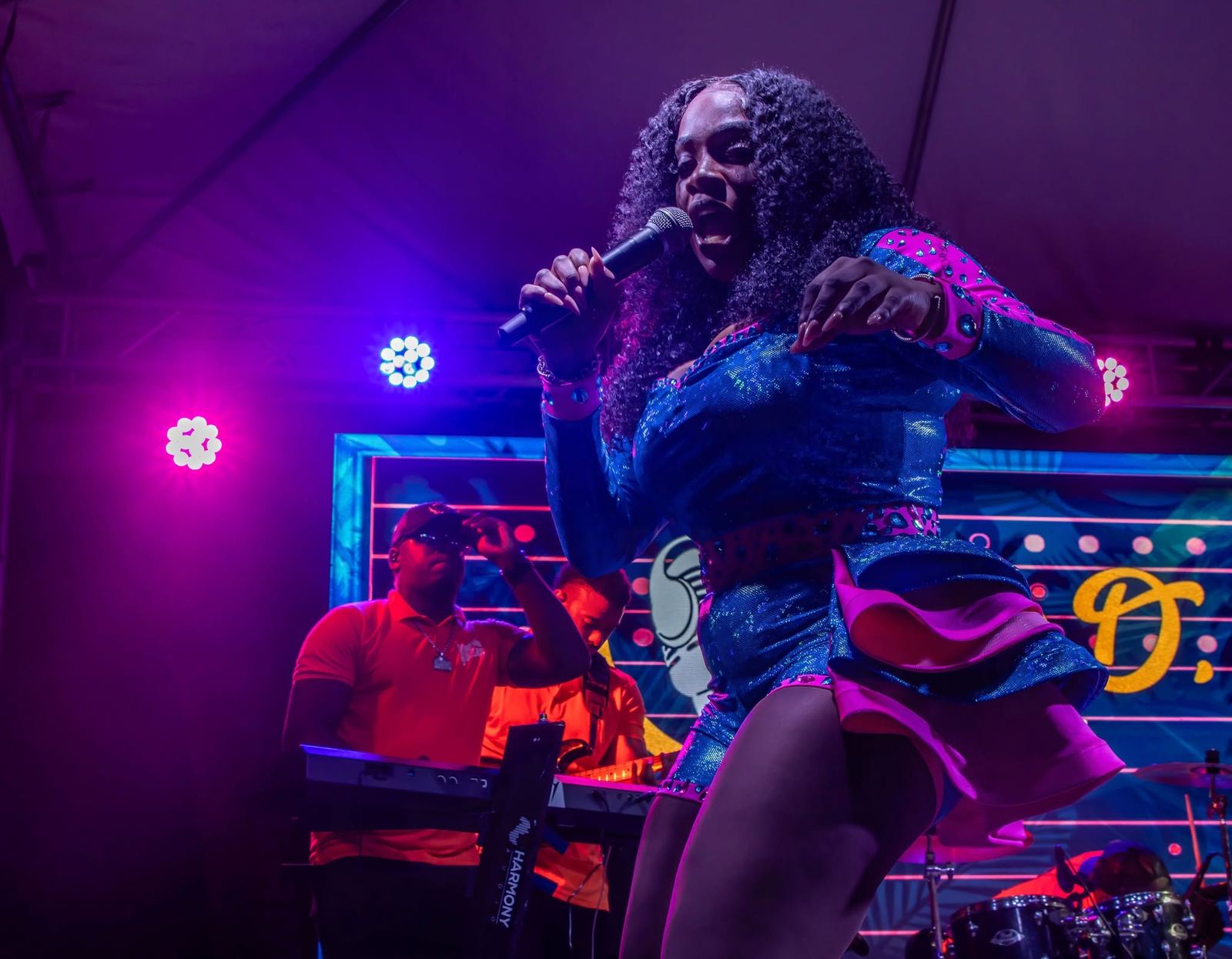 Mama D singing into the microphone with stage lights and her band lit in magenta and blue behind her