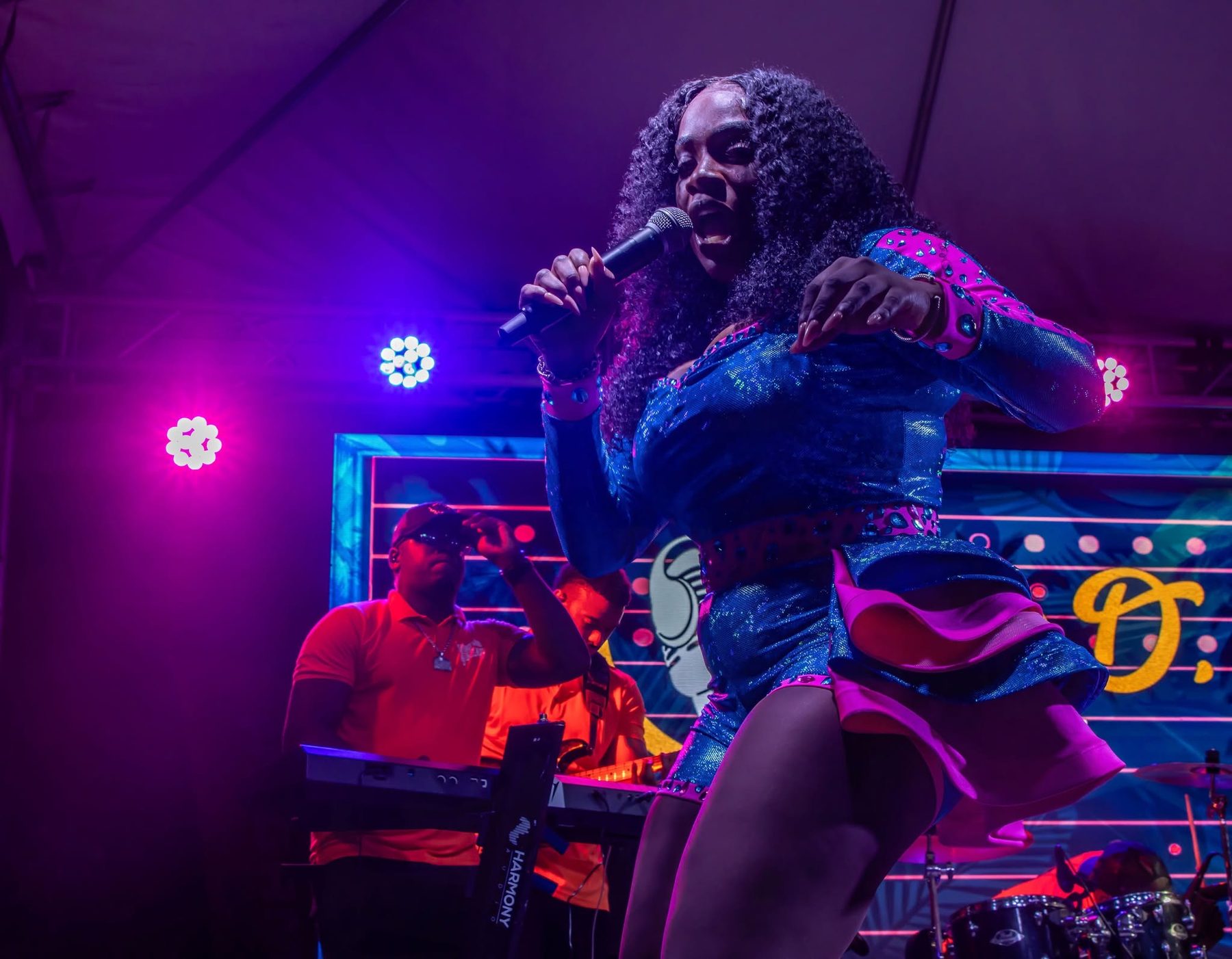 Mama D singing into the microphone with stage lights and her band lit in magenta and blue behind her